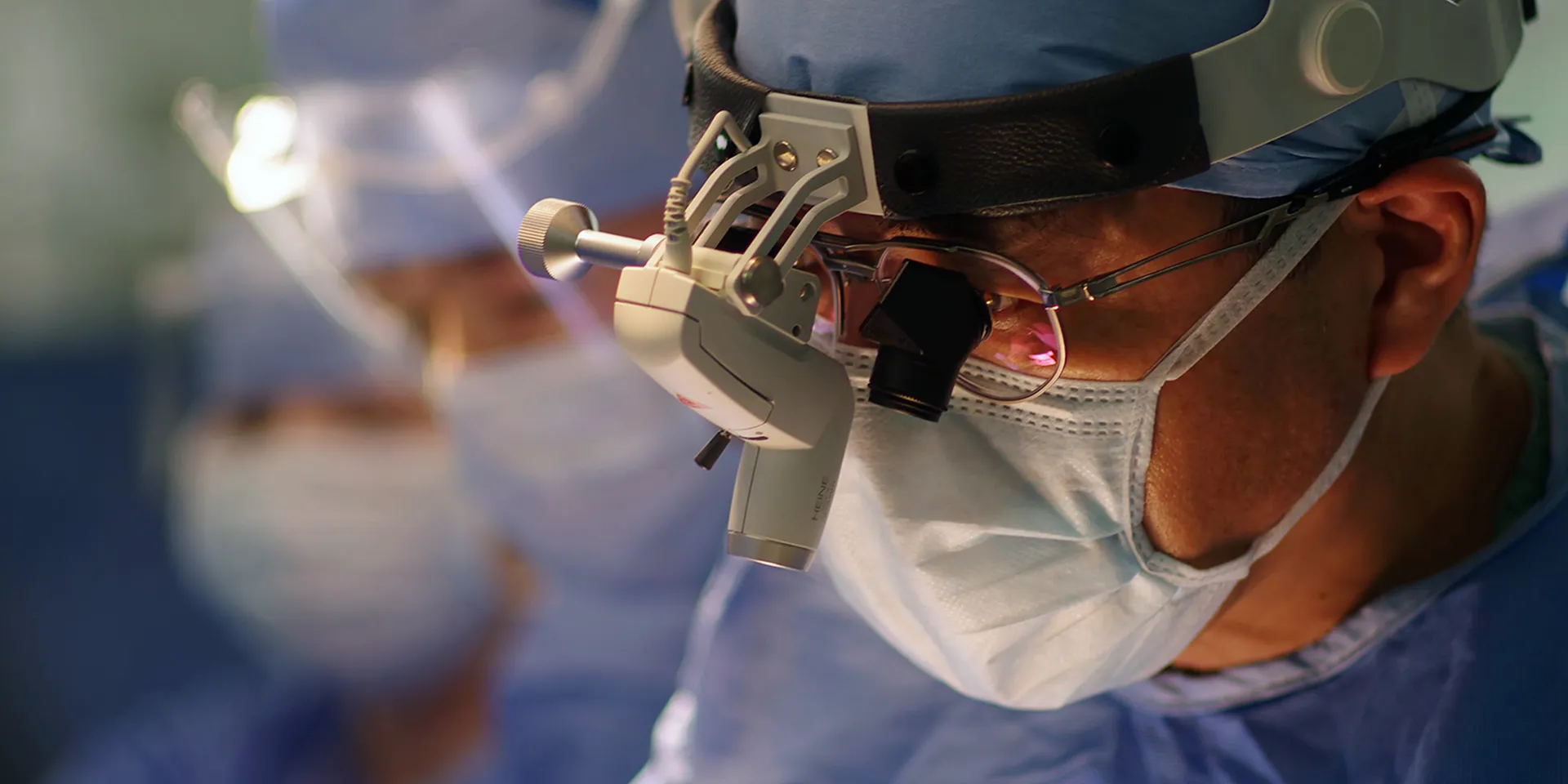 手術中の医師が特別な拡大鏡を装着し、集中している様子。背景には他の医療スタッフが見える。