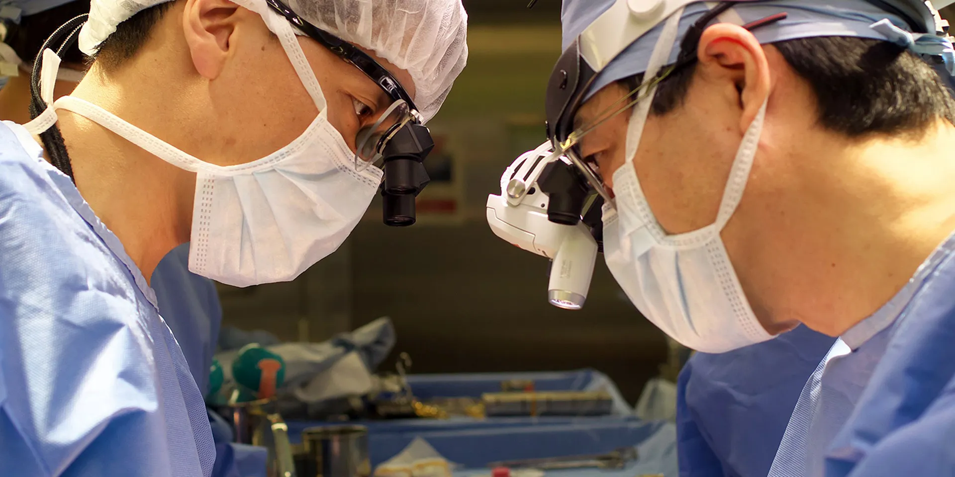 手術中の医師二人が集中している様子。手術用のマスクと帽子を着用し、器具を扱っている。