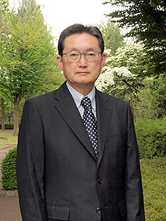 福島県立医科大学の高瀬信弥教授が、緑豊かな背景の中でスーツ姿で立っている写真。