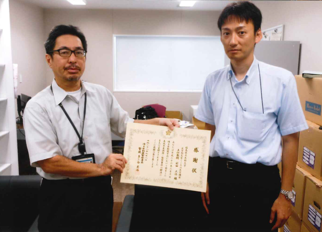 二人の男性が感謝状を持ってオフィスに立っている。背景には棚と箱が見える。男性たちはビジネスカジュアルな服装をしている。