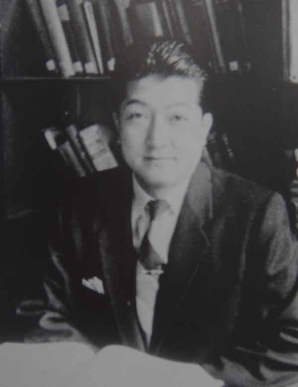 初代教授の飯島進が書籍の前で微笑むモノクロ写真。
