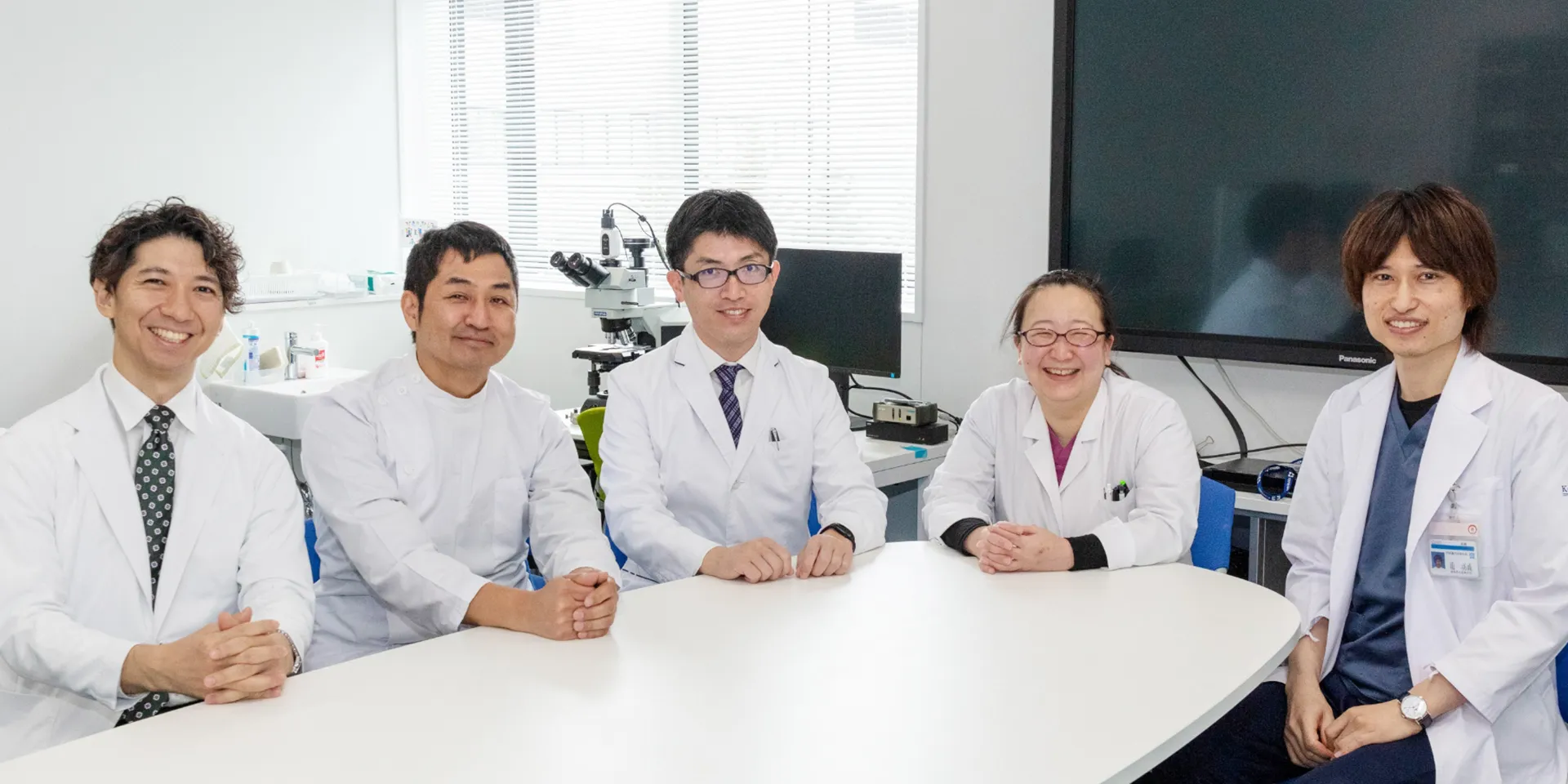 福島県立医科大学の医師たちが白衣を着て、テーブルに座って笑顔でポーズをとっている様子。
