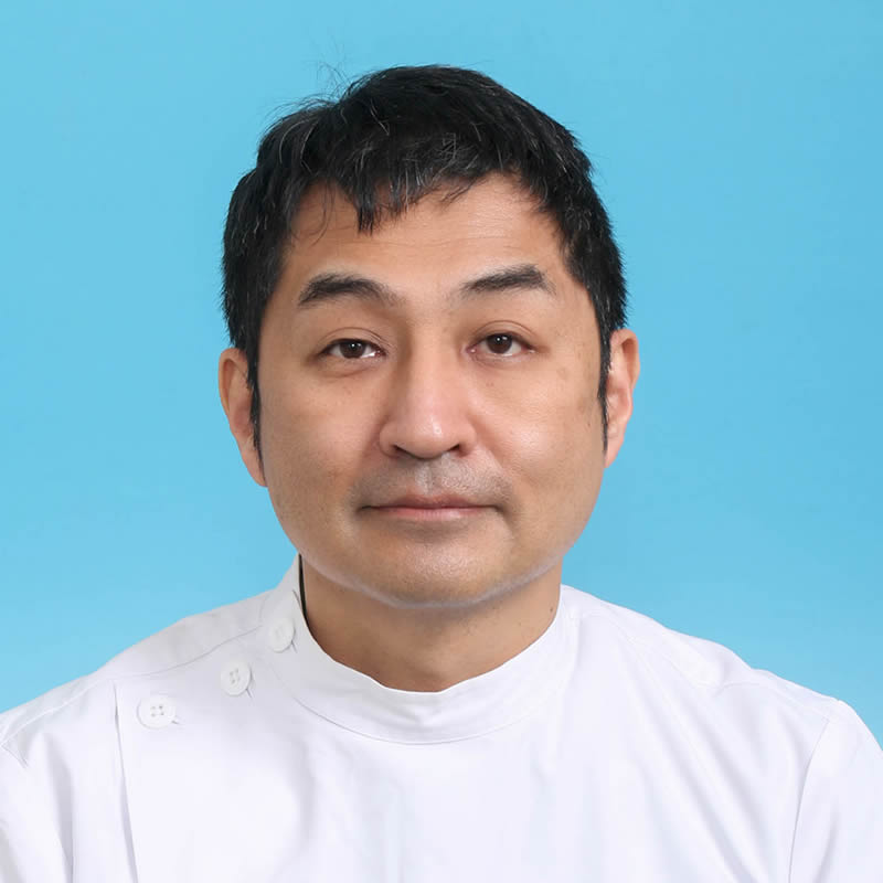 病院助手の中野恵一さんのポートレート。白衣を着て青い背景の前で微笑んでいる。