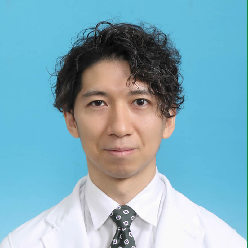 博士研究員の尾崎章彦氏のポートレート。白衣を着用し、青い背景の前で微笑んでいる。