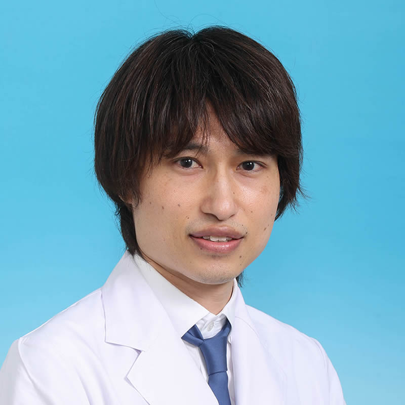 専攻医の塩功貴が白衣を着て微笑むポートレート。背景は青色。