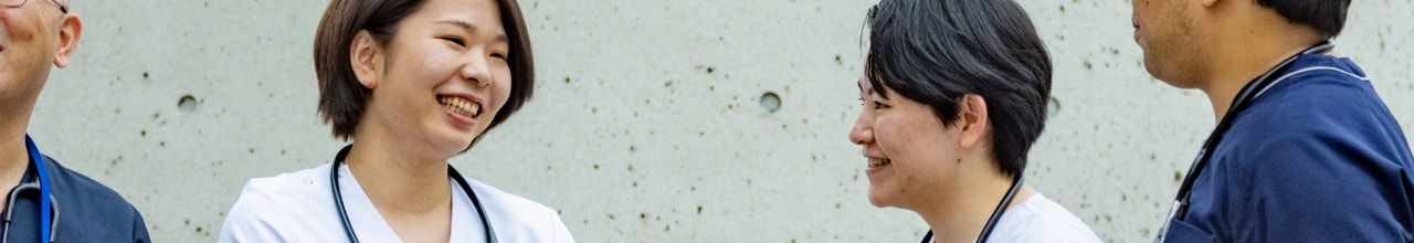 青と白のスクラブを着た4人の医療従事者が、コンクリートの壁を背景に談笑している場面。