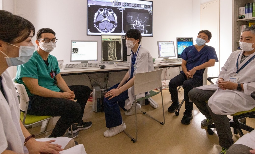 医療従事者がマスクを着用し、コンピュータ画面にはMRI画像が表示されている部屋で会議を行っている。