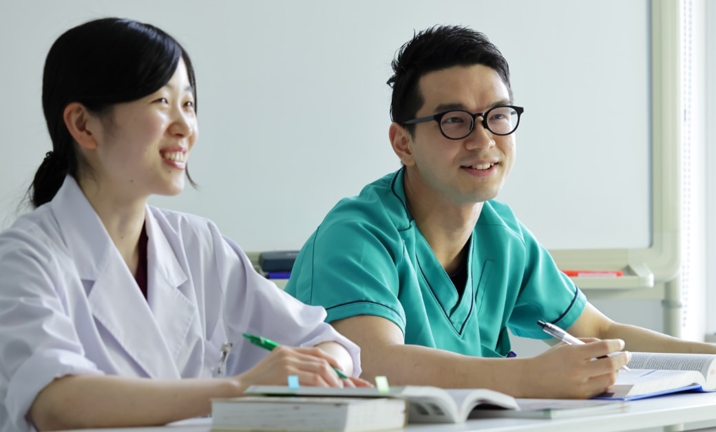 医学生と医療従事者がテーブルで笑顔で勉強している。白衣とスクラブを着て、ペンを持ちノートを前にしている。