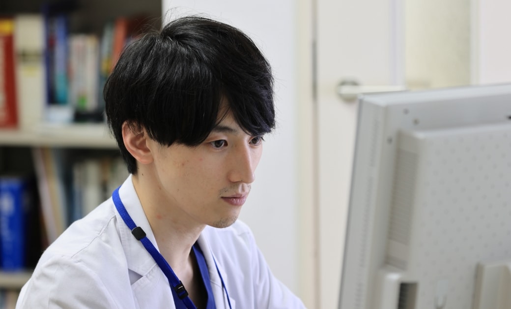 パソコンで作業に集中する白衣を着た若い男性、背景には本棚が見える。