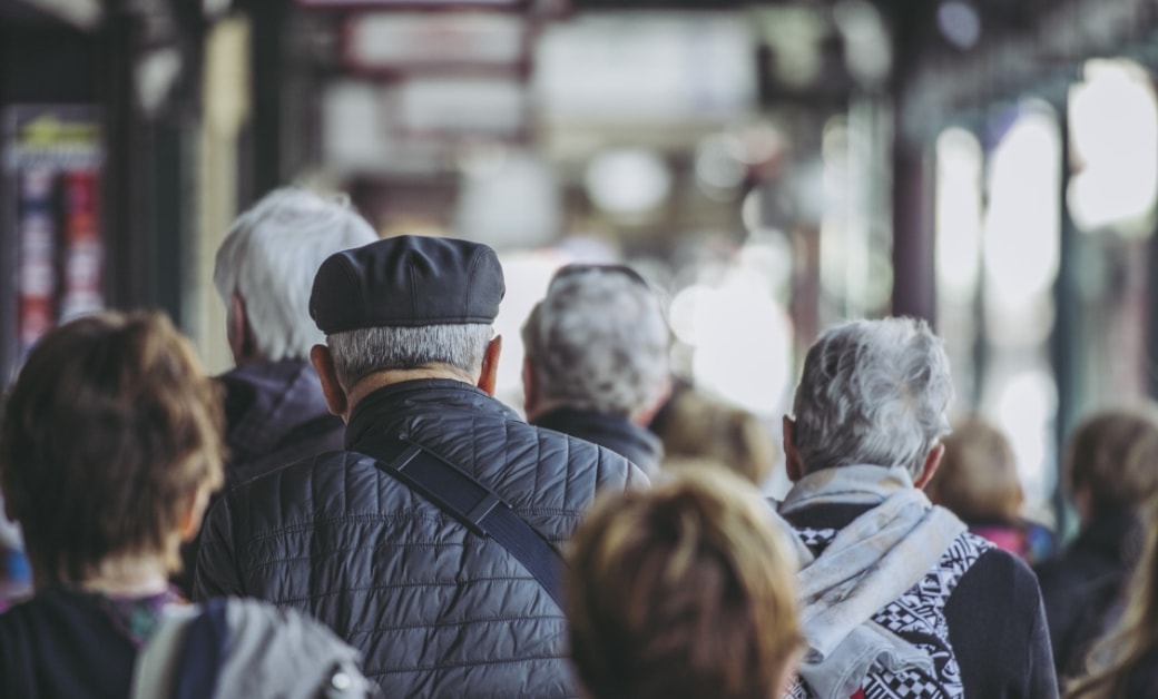 歩道を歩く多くの人々の背中が見えるシーン。年代を感じさせる人々が混雑した街並みを移動中。