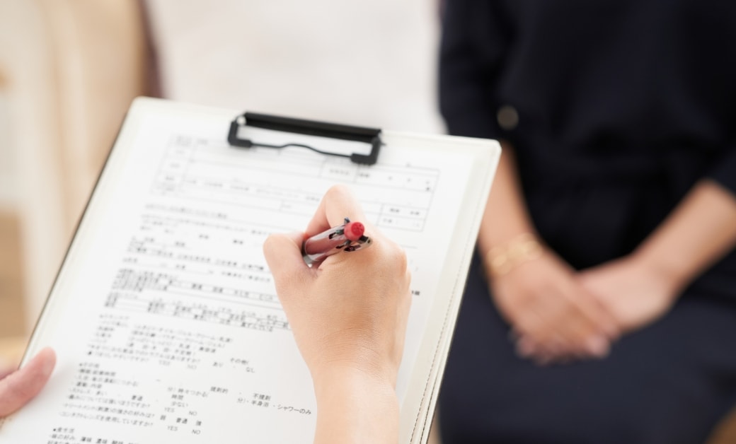 女性がクリップボードに記入し、座っている別の女性が背景にいる医療シーン。