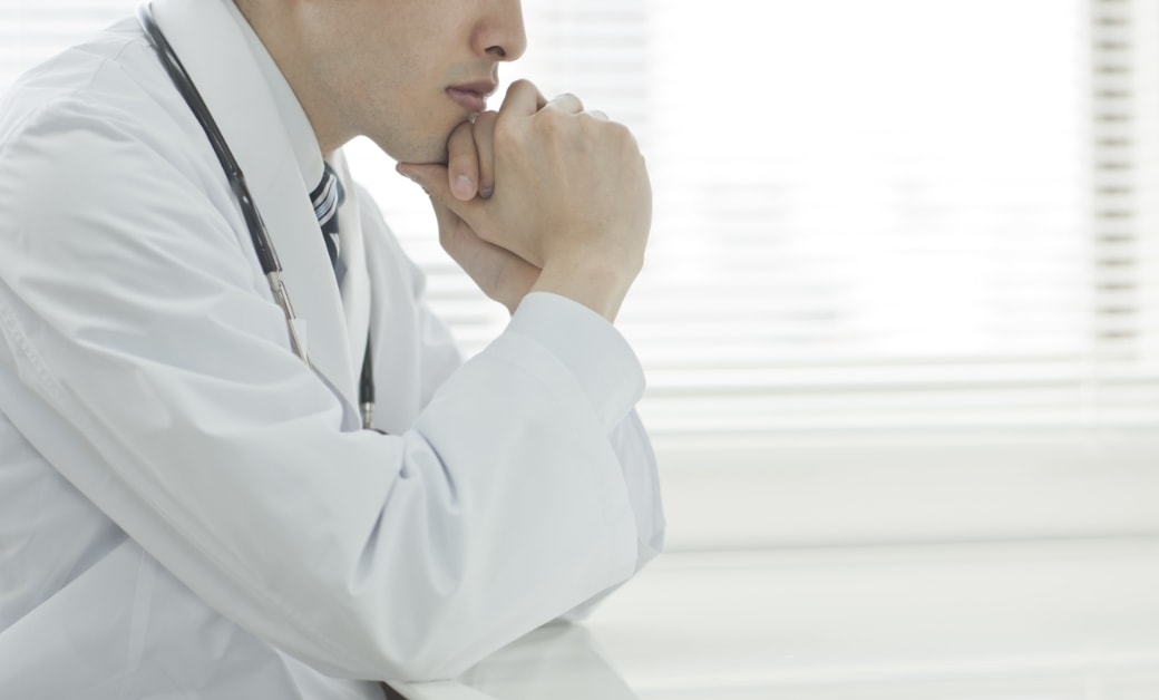 白衣を着た医師が机に肘をつき、手を組んで考え込んでいる姿。背景にはブラインドが見える。