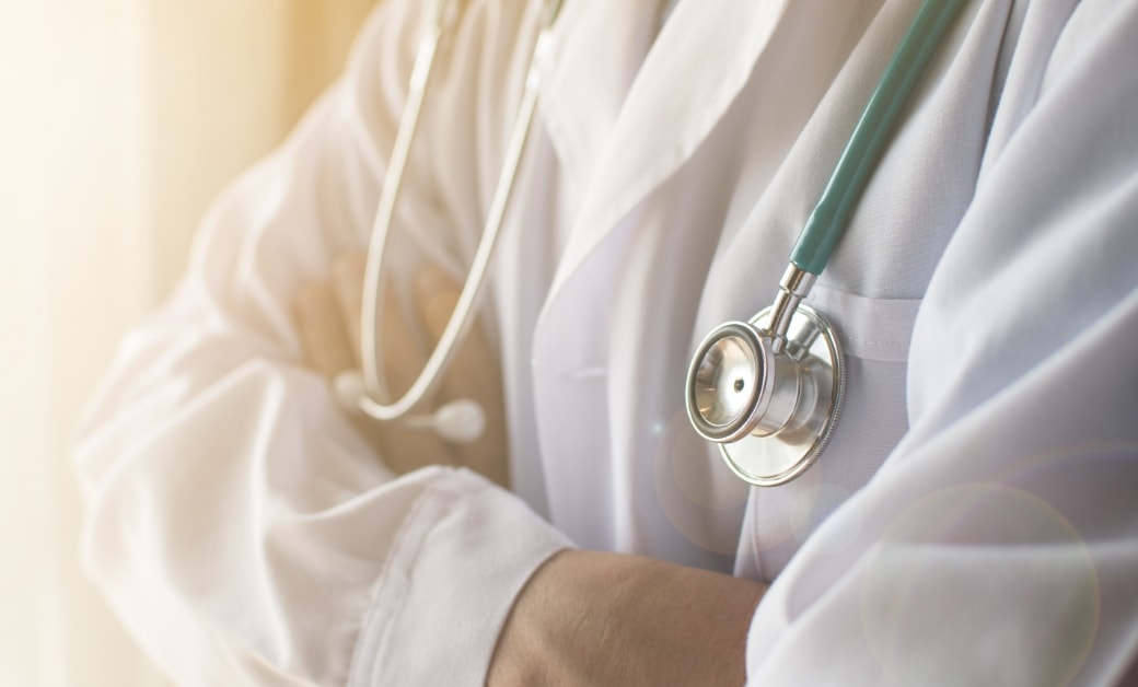 白衣を着た医者が腕を組んでいる。ステートスコープが首にかけられており、柔らかな光が差し込む背景。