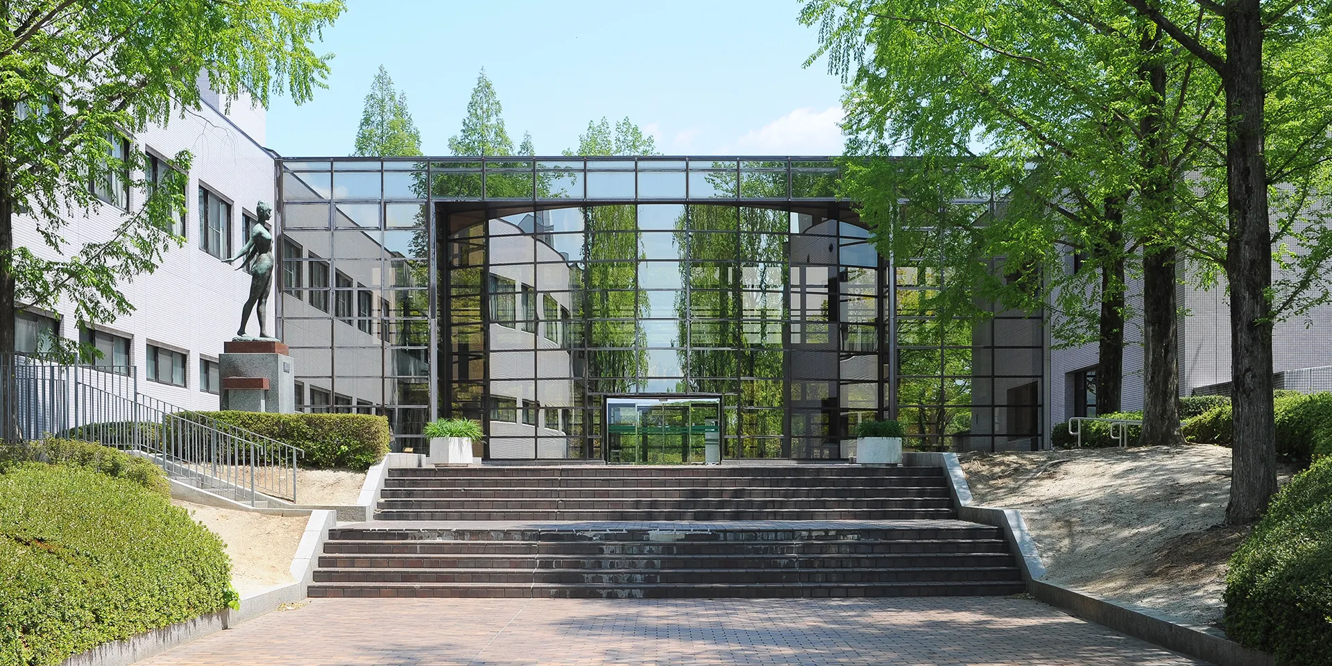 福島県立医科大学のキャンパス入口、緑豊かな木々とガラス張りの建物が特徴的な風景。