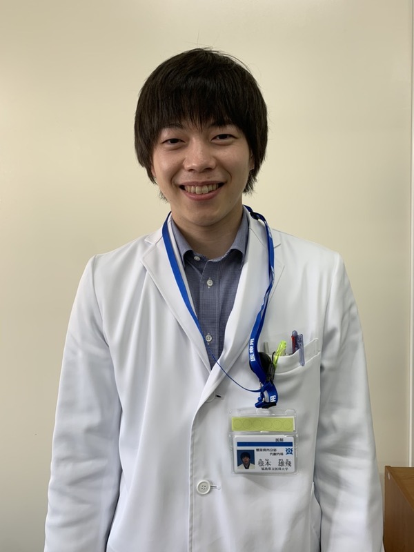 医療白衣を着た根本雄飛さんが笑顔で立っている写真。福島県立医科大学のスタッフ紹介用。