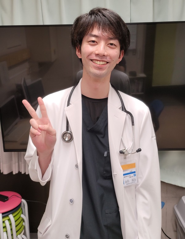 白衣を着た医師が笑顔でピースサインをしている写真。背景にはモニターが映っている。