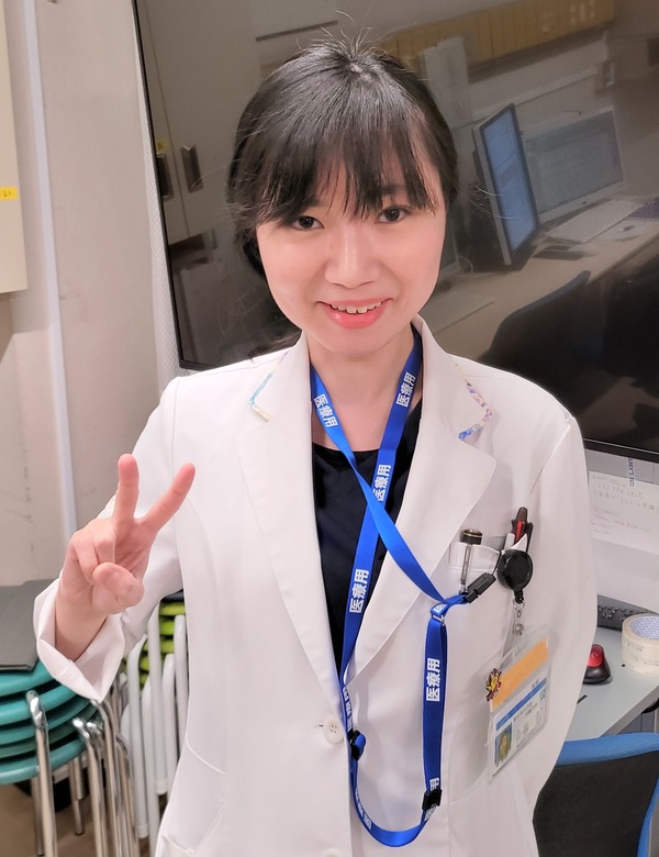 白衣を着た女性医師が笑顔でピースサインをしている写真。背景には医療機器が見える。