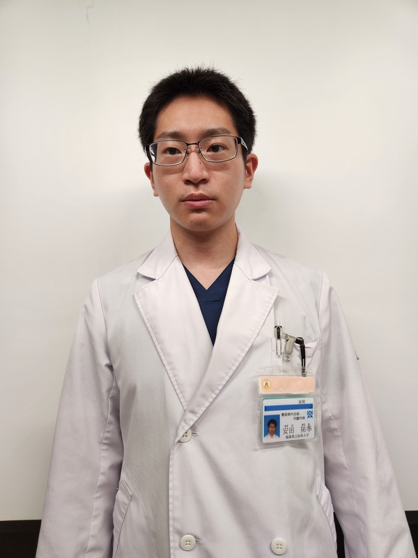白衣を着た男性がカメラに向かって立っている写真。彼は眼鏡をかけ、医療スタッフの名札を付けている。