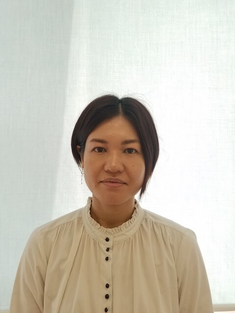 大学院生の城間かおりさんが白いブラウスを着て微笑んでいるポートレート。背景は明るいカーテン。