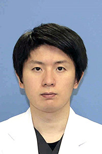 医療スタッフの圓谷秀哲さんのプロフィール写真。白衣を着て、青い背景の前で正面を向いている。
