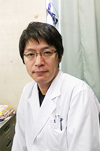 医療白衣を着た男性がカメラに向かって微笑んでいるポートレート。背景には医療機器が見える。