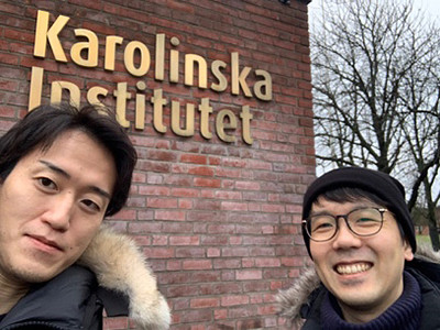 カロリンスカ研究所の看板の前で笑顔の二人が自撮りをしている様子。