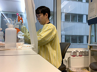 実験室で予防衣を着た研究者が細胞を扱っている様子。窓から外の光が差し込む明るい環境。