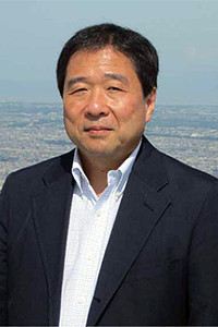 柴田昌彦教授が屋外で微笑んでいるポートレート。背景には都市の風景が広がる。