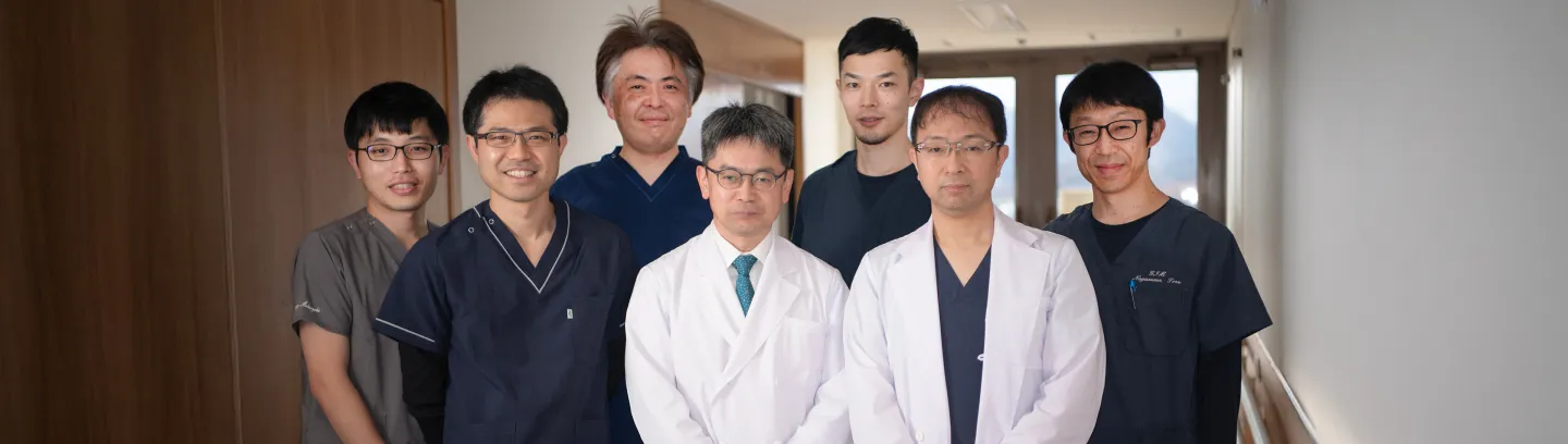 7名の医療従事者が白衣やスクラブを着用し、室内で並んで笑顔でポーズを取っている。背景には明るい廊下が見える。