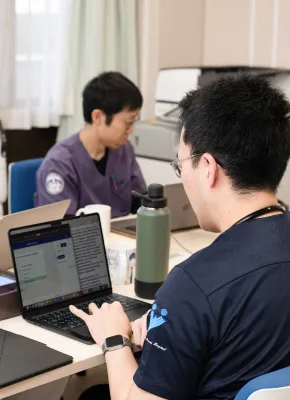 研究室でパソコンを使っている学生たちが、資料を確認しながら作業をしている様子。