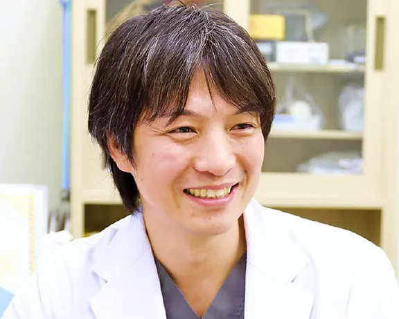 医療現場で微笑む医師が白衣を着ている写真。背景には医療器具が並ぶ棚が見える。