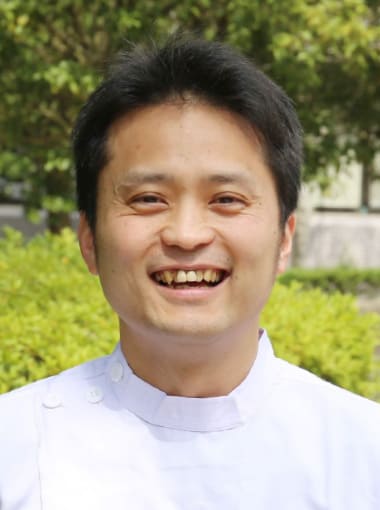 肝胆膵・移植外科学講座の助教、佐藤直哉さんが笑顔で写っている写真。背景には緑の植物が見える。