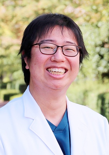 福島県立医科大学の上野智史医師が白衣を着て笑顔でポーズをとっている写真。背景には緑の木々が見える。