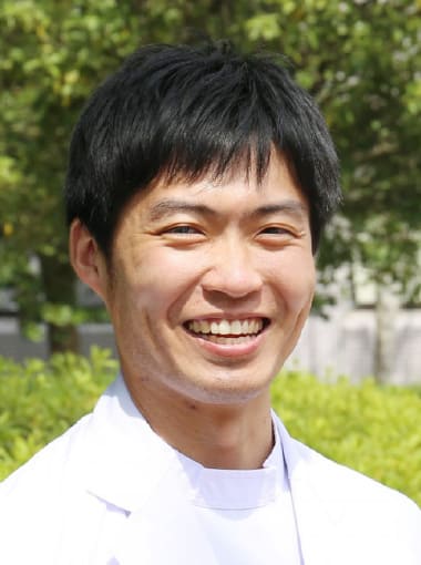 肝胆膵・移植外科学講座の博士研究員、渡邊淳一郎さんが笑顔でポーズをとっている写真。背景には緑の植物が見える。