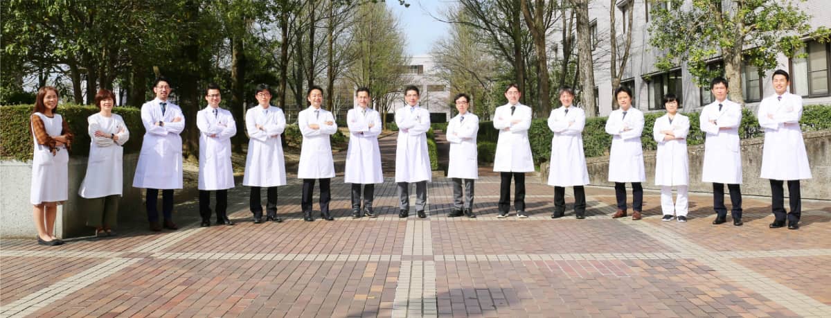 福島県立医科大学のスタッフが並んで立っている写真。医療従事者の白衣姿で、背景には緑の木々が見える。