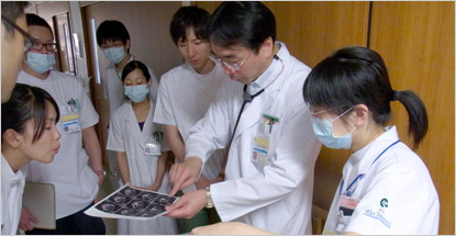 医師が医療スタッフに画像を見せながら説明しているシーン。周囲には数人の医療従事者が集まっている。
