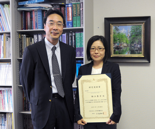 物江恭子先生が日本肝臓学会研究奨励賞を受賞し、賞状を持っている写真。背景には書籍棚と絵画が見える。