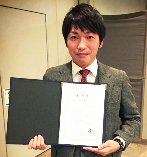 岡井研先生が第1回G-PLUS優秀賞を受賞し、賞状を手に微笑んでいる姿。