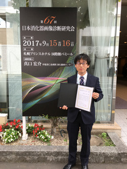 第67回日本消化器画像診断研究会で優秀ポスター賞を受賞した浅間宏之先生が、賞状を持って笑顔で立っている写真。背景には会議のポスターが掲示されている。