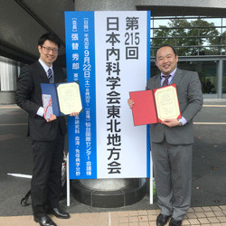 第215回日本内科学会東北地方会の前で、受賞した佐藤秀紀先生と郡司直彦先生が賞状を持って笑顔で立っている様子。