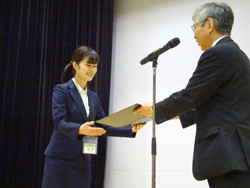 受賞式で表彰状を受け取る女性と、彼女に授与する男性の様子。