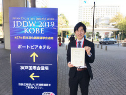 第27回日本消化器関連学会週間で若手奨励賞を受賞した杉本充先生が、神戸の会場前で賞状を持っている様子。