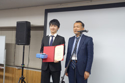 受賞式で表彰状を持つ学生と講師が並んでいる様子。
