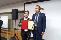 受賞式で表彰状を持つ二人の男性が写っている。背景にはスピーカーとスクリーンがある。