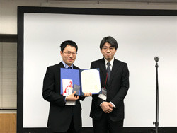 受賞式で表彰状を持つ二人の男性が写っている写真。