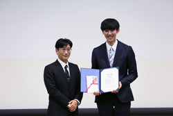 受賞式で表彰状を持つ二人の男性が写っている写真。左側の男性はスーツを着ており、右側の男性は笑顔で表彰状を掲げている。