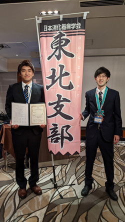 受賞を祝う場面で、東北支部の横断幕の前に立つ二人の学生が、賞状を持っている。