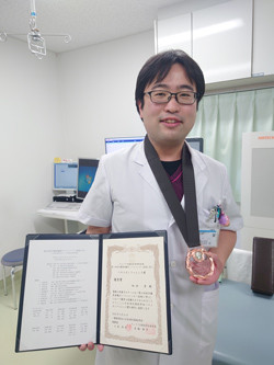 受賞した医師が賞状とメダルを持って微笑んでいる写真。背景には医療機器が見える。