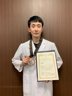受賞した医師が白衣を着て、メダルと賞状を手に持っている写真。背景は木目調の壁。