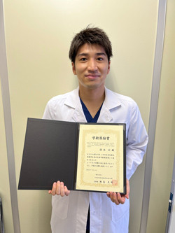 医療白衣を着た男性が、受賞証書を持って微笑んでいる写真。背景は無地の壁。